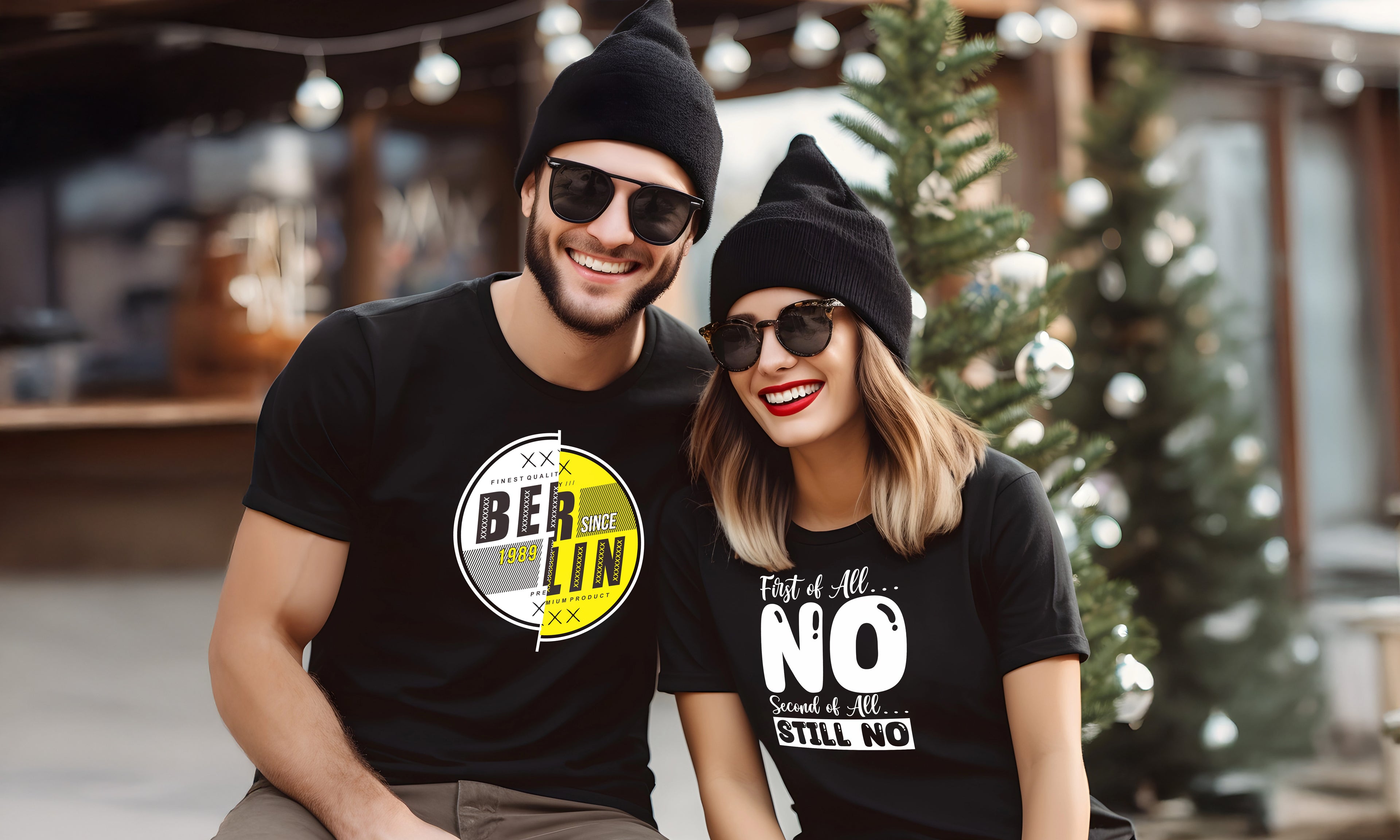 Ein Mann und eine Frau mit schwarzen Beanies und Sonnenbrillen sitzen lächelnd draußen. Beide tragen schwarze T-Shirts mit auffälligen grafischen Mustern. Im Hintergrund steht ein kleiner immergrüner Baum, der mit festlichen Lichtern geschmückt ist.