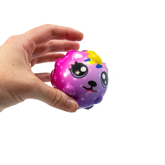 Eine Hand hält ein kleines, rundes, buntes Spielzeug mit einem süßen Gesicht. Der Animal High Bounce Ball von www.buyon.at ist überwiegend rosa und lila, hat große schwarze Augen, eine kleine schwarze Nase und einen lächelnden Mund, aus dem eine winzige rosa Zunge heraushängt.