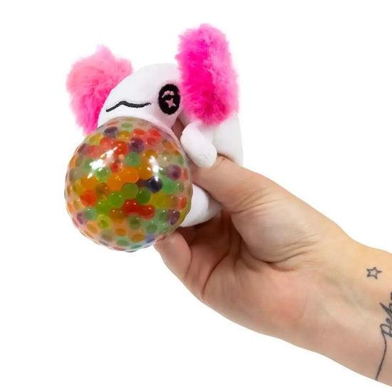 Eine Hand hält einen weißen Axolotl Pearl Squish Ball von www.buyon.at mit rosa flauschigen Ohren und einem schwarz genähten Auge. Das Spielzeug umklammert einen großen, transparenten Ball, der mit bunten Gelperlen gefüllt ist und sich ideal zur sensorischen Stimulation eignet. Auf dem schlichten weißen Hintergrund ist in der Nähe des Handgelenks ein kleines Sterntattoo zu sehen.