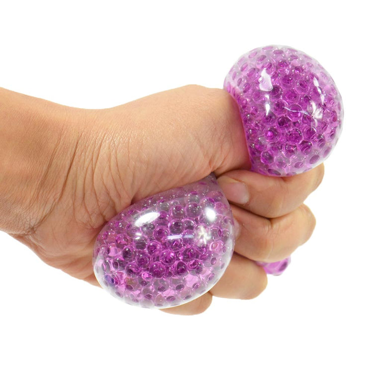 Eine Hand drückt den 7 cm großen Crystal Beads Ball von www.buyon.at und gibt kleine, violette Perlen im Inneren frei, die ihn unter Druck wölben und dehnen. Die transparente Außenseite ermöglicht es, durch die Perlen hindurchzusehen, was ein strukturiertes und optisch anregendes Erscheinungsbild bietet, das perfekt zum Stressabbau geeignet ist.