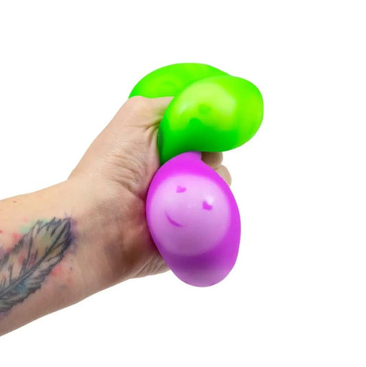 Eine Hand mit einer Federtätowierung drückt den Emotions Squish Ball von www.buyon.at. Der violette Teil ist mit einem schlichten Smiley-Design versehen. Der Stressball wirkt weich und flexibel und sorgt vor einem schlichten weißen Hintergrund für sensorische Stimulation.