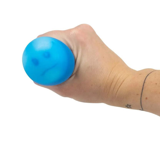 Eine Hand hält einen blauen Emotions Squish Ball von www.buyon.at mit einem darauf gezeichneten traurigen Gesicht. Die Person hat ein kleines Stern-Tattoo am Handgelenk und ein zusätzliches Linien-Tattoo weiter oben am Unterarm. Der Hintergrund ist schlicht weiß und hebt den Ball hervor, der zur sensorischen Stimulation gedacht ist.