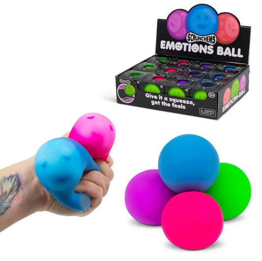 Eine Hand drückt einen blauen Emotions Squish Ball von www.buyon.at, auf dem ein lächelndes Gesicht abgebildet ist. Darunter sind vier bunte Stressbälle in den Farben Blau, Rosa, Grün und Lila aufeinander gestapelt. Im Hintergrund ist eine Displaybox mit weiteren Emotions Squish Balls zu sehen, die mit „sensorische Stimulation“ beschriftet ist.