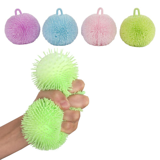 Eine Hand zerquetscht zwei grüne, stachelige Fluffy Quetschball Pastells von www.buyon.at. Über der Hand befinden sich vier weitere Stressbälle in verschiedenen Farben: lila, blau, rosa und gelbgrün. Jeder 9 cm große Ball hat oben einen Schlaufengriff. Ideal zur Entspannung und zur Verbesserung der Feinmotorik.