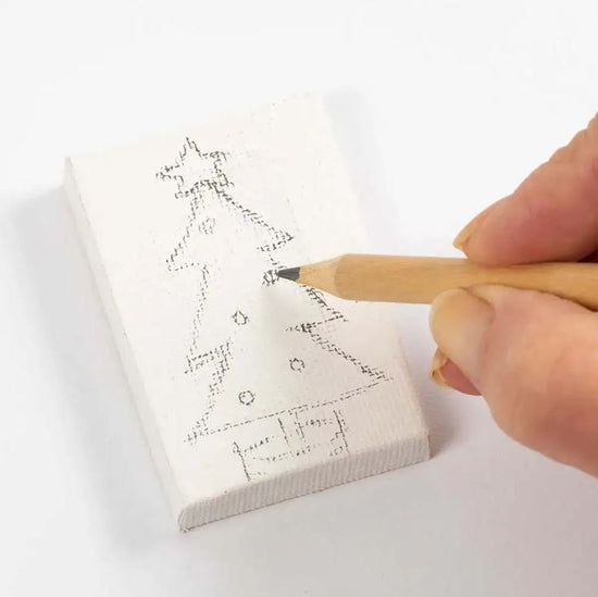 Nahaufnahme einer Hand, die einen Bleistift hält und auf einer kleinen weißen Leinwand aus dem Malerei, Mini-Kreativ-Set von www.buyon.at die gepunkteten Umrisse eines Weihnachtsbaums skizziert. Der Baum hat einen Stern an der Spitze und Ornamente an den Zweigen.