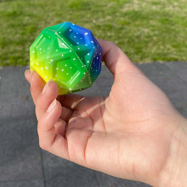 Eine Hand hält einen bunten Mega High-Bounce Ball, GEO von www.buyon.at. Der Ball hat die Form eines Dodekaeders mit Farbverläufen von Blau über Grün zu Gelb und weist auf seinen Facetten ein weißes Punktmuster auf. Der Hintergrund zeigt einen Bürgersteig und etwas Gras in einer Außenumgebung.