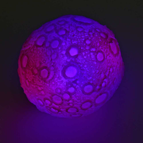 Ein Mondball, Bounce Ball mit LED von www.buyon.at, mit einer kraterartigen Oberfläche, die an den Mond erinnert und von violetten und rosa Lichtern beleuchtet wird, liegt vor einem dunklen Hintergrund. Dieses himmlisch inspirierte Zappelspielzeug ist sowohl faszinierend als auch beruhigend anzusehen.