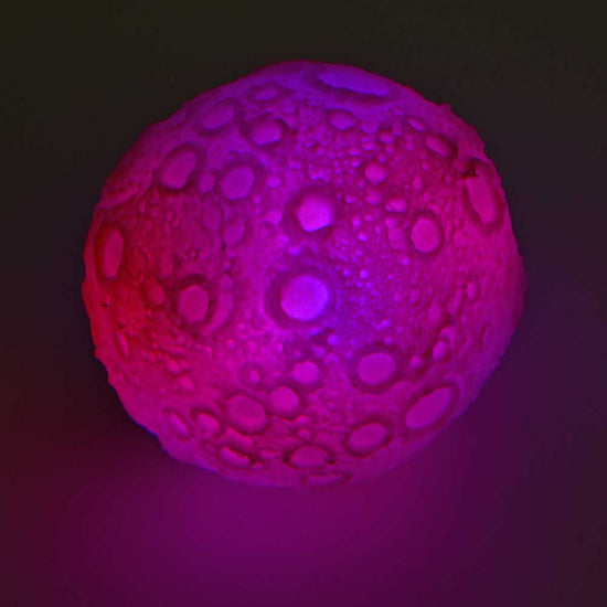 Ein Mondball von www.buyon.at, bekannt als Bounce Ball mit LED, ähnelt einem stilisierten Mond mit kraterartigen Vertiefungen, der mit einem kräftigen rosa und violetten Lichtverlauf vor einem dunklen Hintergrund beleuchtet wird.