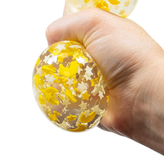 Eine Hand drückt einen klaren, runden Moons & Stars Squish Ball von www.buyon.at, der mit gelben Halbmonden und weißen Sternen gefüllt ist. Der flexible Stressabbau-Ball wird mit der Hand zusammengedrückt, wodurch die Verzierungen im Inneren sichtbar werden und er als beruhigendes Zappelelement dient.