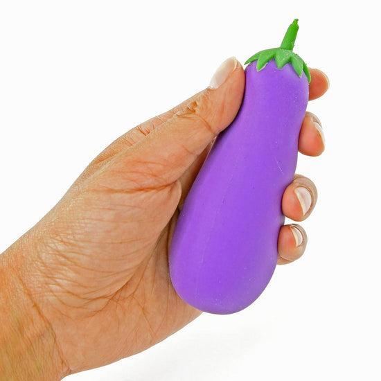 Eine Hand hält einen lila Stressball in Form einer Aubergine, bekannt als Quetsch Aubergine von www.buyon.at. Das Spielzeug, das an der Spitze einen grünen Stiel aufweist, wird vor dem weißen Hintergrund sanft von der Hand gedrückt und dient als hervorragendes Stressabbau- und Spaßspielzeug für Kinder.