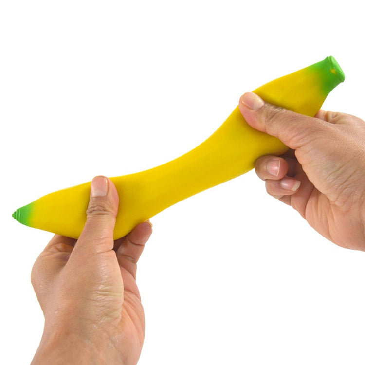 Eine Person streckt ein leuchtend gelbes, bananenförmiges Stressspielzeug namens Squeeze Banane von www.buyon.at, das an beiden Enden grüne Spitzen hat. Sie greift das Spielzeug fest mit beiden Händen; die linke Hand hält ein Ende, während die rechte Hand am anderen zieht, um es zum Stressabbau zu dehnen.