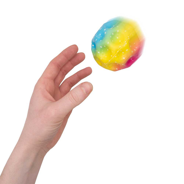 Eine Hand wirft einen Rainbow Mega High-Bounce Ball von www.buyon.at in die Luft. Der Ball mit Regenbogenverlauf und Glitzer erzeugt vor einem schlichten weißen Hintergrund einen skurrilen Effekt.