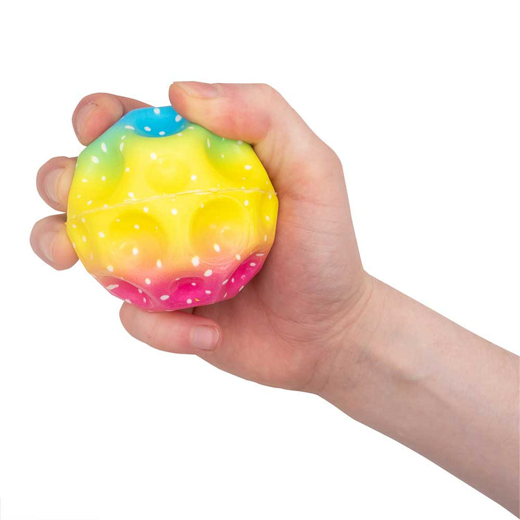 Eine Hand drückt einen bunten Rainbow Mega High-Bounce Ball von www.buyon.at, der einen Farbverlauf in Gelb, Grün, Blau und Rosa mit weißen Punkten aufweist. Die Oberfläche des Balls ist mit kleinen Unebenheiten strukturiert, die ein taktiles Gefühl vermitteln.