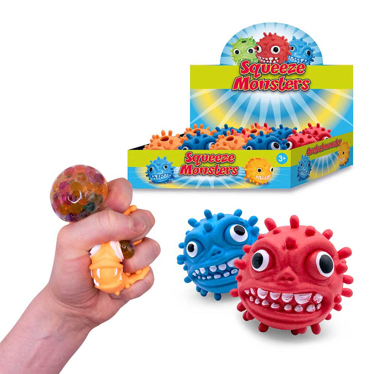 Eine Hand drückt ein orangefarbenes „Squeeze Monster Face“-Spielzeug mit Wasserperlen darin. Im Hintergrund ist eine Schaubox mit roten, blauen und grünen stacheligen Monsterspielzeugen mit der Aufschrift „www.buyon.at Squeeze Monsters“ zu sehen. Im Vordergrund sind zwei größere Monsterspielzeuge, ein blaues und ein rotes, zu sehen. Perfekt zum Stressabbau!