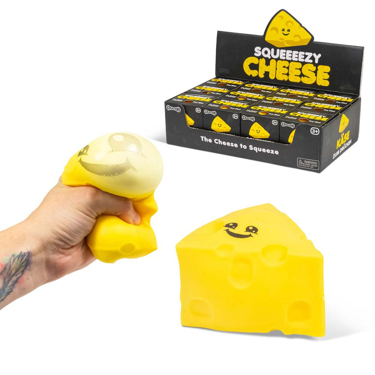 Eine Hand drückt ein gelbes, käseförmiges Stressspielzeug mit einem lächelnden Gesicht. Unten ist der Squeezy Cheese von www.buyon.at in seiner Originalform zu sehen, mit weiteren Spielzeugen in einer Hintergrundbox mit der Aufschrift „Squeezy Cheese“. Dieses Stressabbau-Tool hat eine beruhigende Wirkung und ist perfekt zum Entspannen.