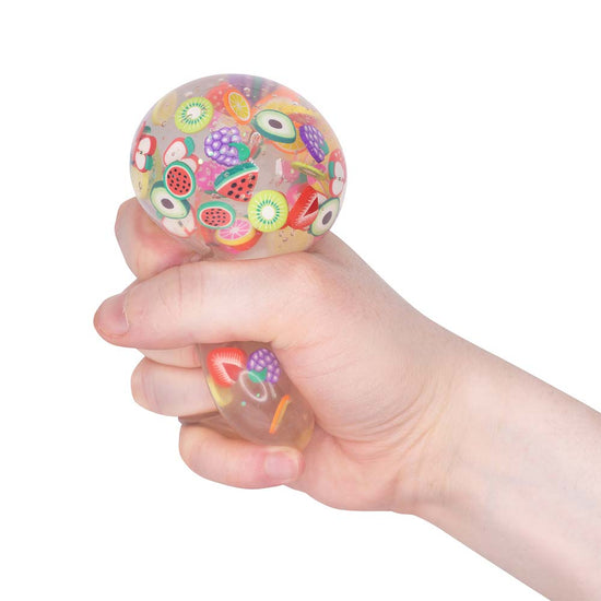 Eine Hand drückt ein durchsichtiges, rundes, weiches Spielzeug namens Tutti Frutti Squish Ball von www.buyon.at. Das Spielzeug ist transparent und mit kleinen, bunten, fruchtförmigen Stücken gefüllt, die an Wassermelone, Kiwi, Avocado und andere Früchte erinnern – perfekt zum Entspannen und Stressabbau.