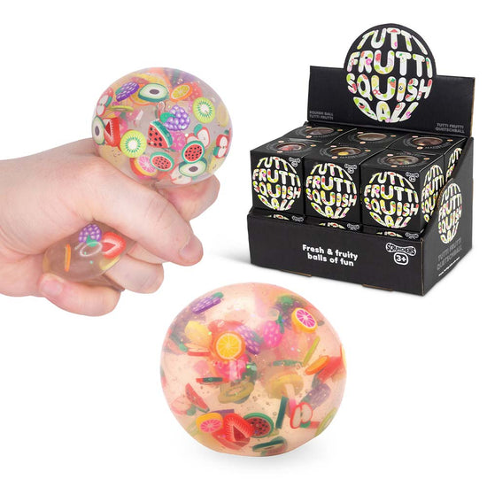 Eine Hand drückt einen transparenten, weichen Tutti Frutti Squish Ball von www.buyon.at, der mit bunten, kleinen Fruchtstücken gefüllt ist und sich hervorragend zum Stressabbau eignet. Darunter ist ein weiterer Squish Ball zu sehen. Im Hintergrund ist eine schwarze Box mit der Aufschrift „Tutti Frutti Squish Ball“ zu sehen, in der sich mehrere Squish Bälle befinden und die unendliche Entspannung bieten.